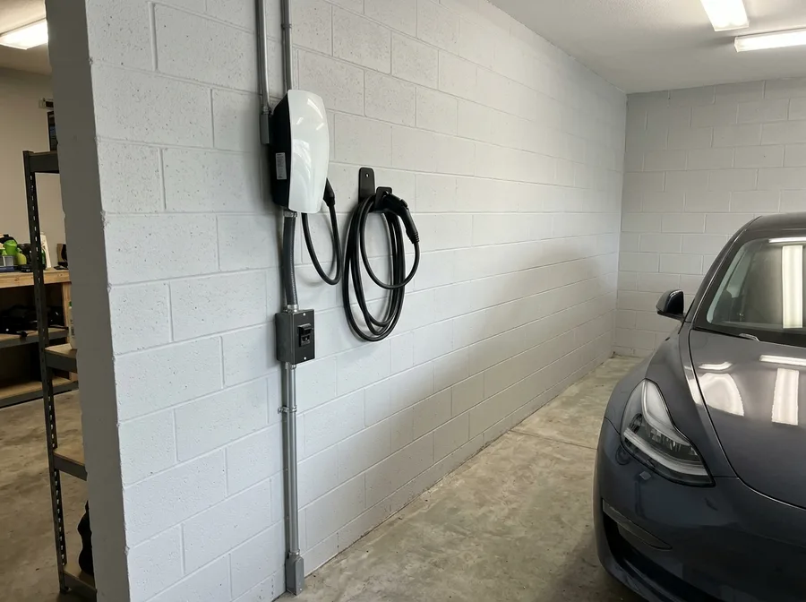 A finished EV charger mounted on a garage wall with its whip and conduit run cleanly down to a connected disconnect box, cord holstered in the hanger, the nose of a parked car at the edge of frame