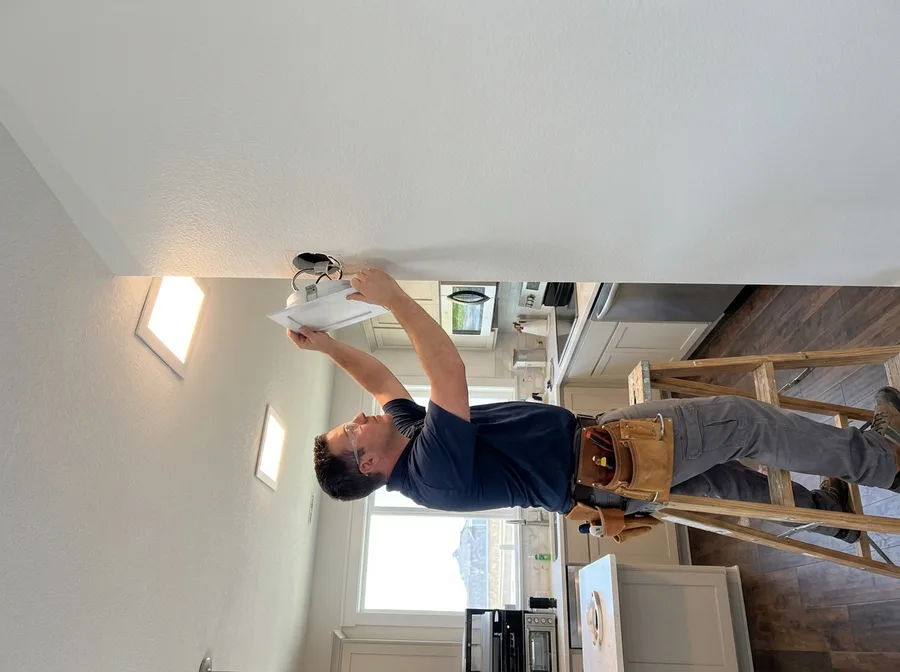 Looking up from below at an electrician on a step ladder twisting the trim of a flush ceiling fixture into its installed mounting bracket in a bright kitchen, a second finished fixture lit further down the ceiling